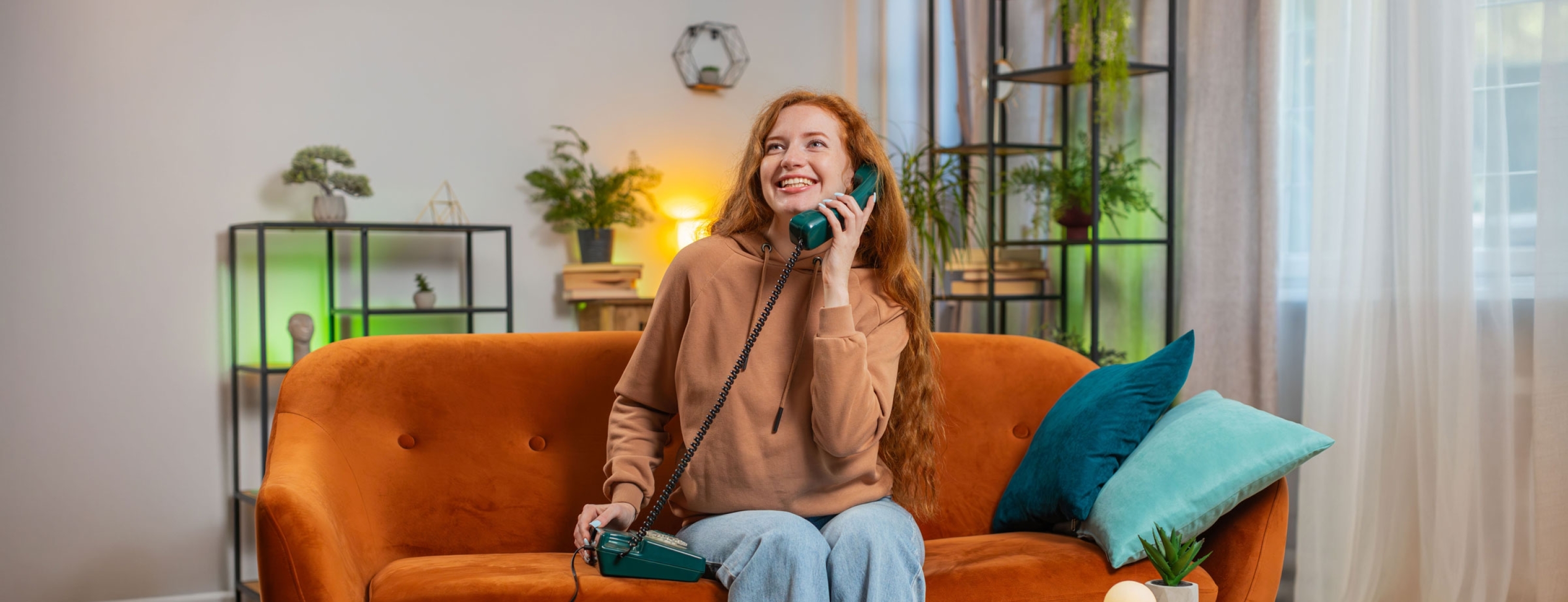 Smiling young woman making wired telephone call conversation with friends sitting on couch at home. Happy excited redhead girl enjoying old-fashioned retro phone from 90s talking in living room.