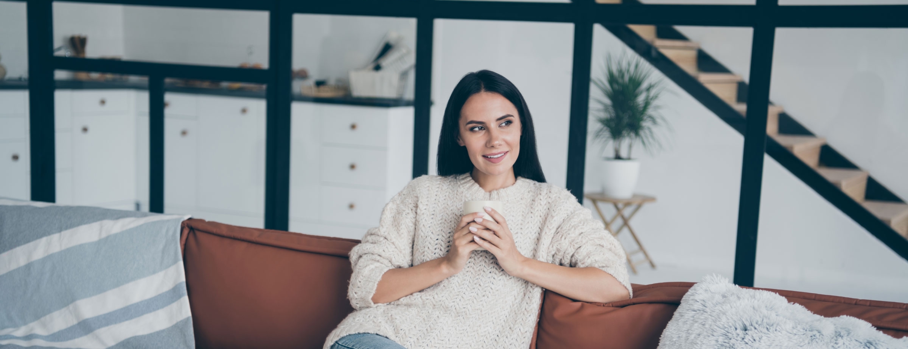 Positive cheerful woman sit on brown leather divan hold cup with latte cappuccino stare window feel inspired imagination dreamy in house indoors